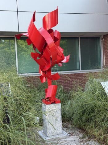 A tall abstract outdoor sculpture by Richard Pitts composed of twisting, ribbon-like forms painted in bright red, rising upward in dynamic curves. The sculpture is mounted on a silver, textured pedestal and set among tall grasses in front of a modern building with brick and metal siding.